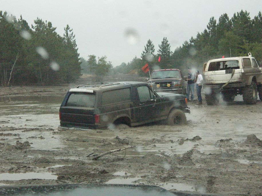 Black Ford Bronco stuck in mud, 3/4 front view. Other trucks visible in background