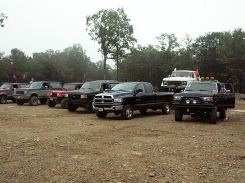 1995 Ford Bronco - A row of lifted 4x4s parked on a gravel staging lot at an off-road park on an overcast day, including a black Dodge Ram quad cab, several o…