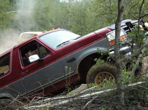 1995 Ford Bronco - A maroon-over-gray Ford Bronco pitched nose-up climbing a dirt bank in the brush, throwing dust as the driver looks out the open window. Vi…