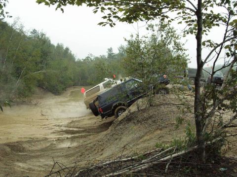 1995 Ford Bronco - Two SUVs tackle a challenging muddy off-road course, kicking up dirt as they ascend a steep incline.