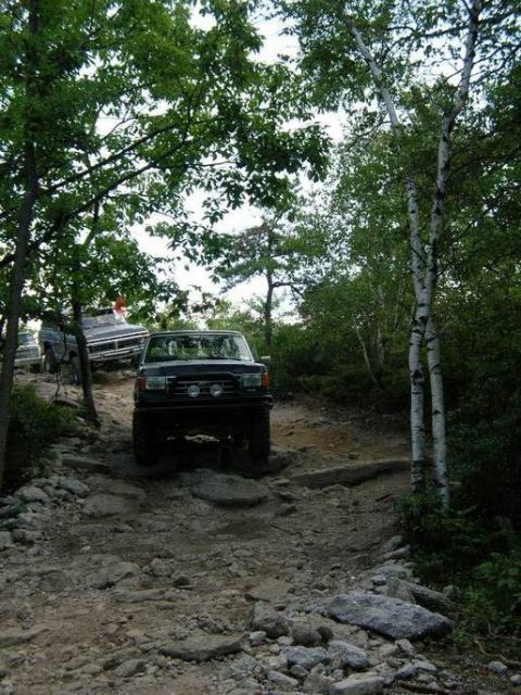 1995 Ford Bronco - Two SUVs navigate a challenging, rocky off-road trail surrounded by trees.