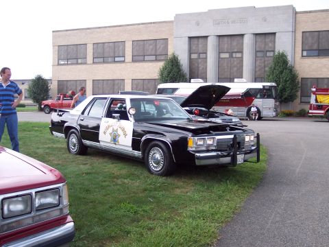 2001 Ford Crown Victoria - A black and white police car is parked on the grass with its hood open, alongside other vehicles and a building.