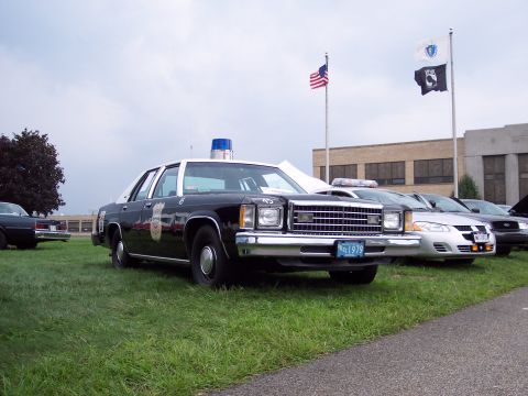 2001 Ford Crown Victoria - This police car, marked with a 1979 license plate, is parked on grass with other vehicles and flags in the background.