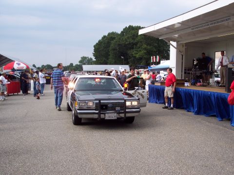 2001 Ford Crown Victoria - A police car with its lights on is parked in front of a stage at an outdoor event.