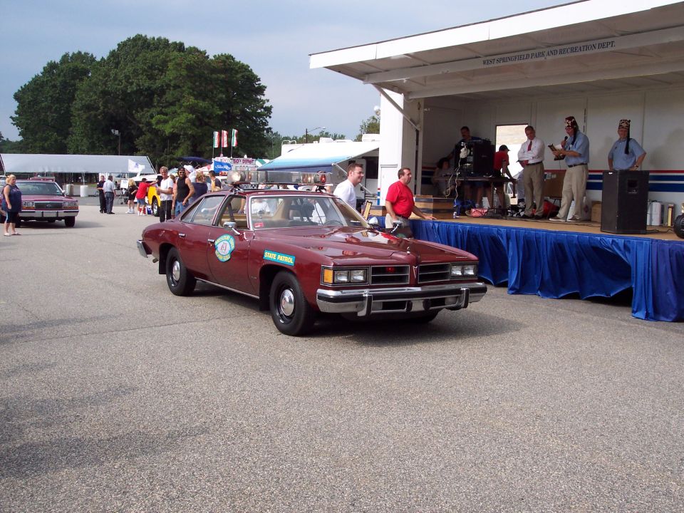 Maroon state patrol car with lights on top, driving past a stage where people are standing