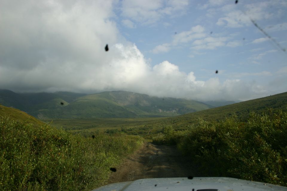 White Jeep hood on a dirt road with green hills and cloudy sky