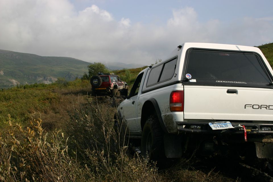 White Ford pickup with camper shell follows a red Jeep on a muddy off-road trail through green hills