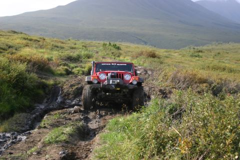 2003 Jeep TJ - A red Jeep TJ navigates a muddy trail on a grassy hillside, with a mountain range visible in the distance.