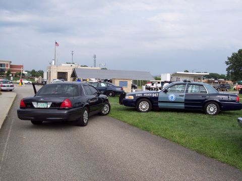 2001 Ford Crown Victoria - Two police vehicles, a dark Ford Crown Victoria and a marked State Police car, are parked side-by-side at an outdoor event.