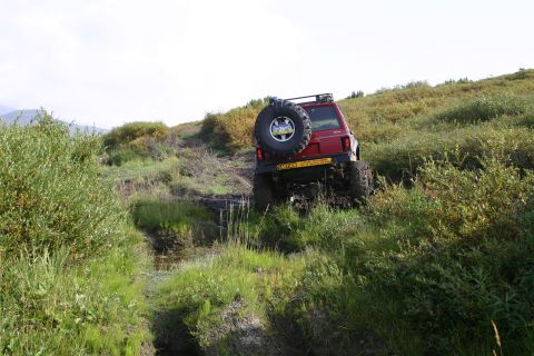 2003 Jeep TJ - A red Jeep Cherokee XJ navigates a challenging off-road trail, kicking up mud as it ascends a steep incline.
