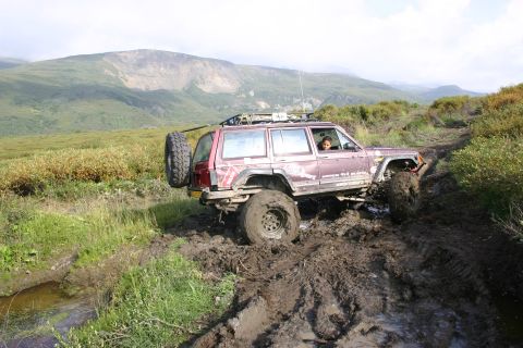 2003 Jeep TJ - A maroon Jeep Cherokee XJ navigates a challenging muddy trail in a mountainous, grassy landscape.