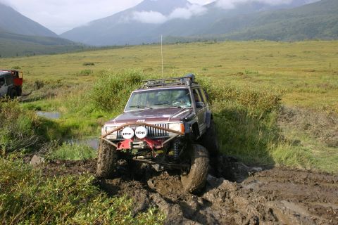2003 Jeep TJ - A maroon Jeep Cherokee XJ struggles through a muddy trail with mountains in the distance; a second Jeep is visible in the background.