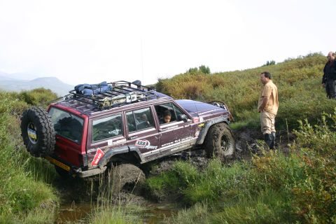 2003 Jeep TJ - A maroon Jeep Cherokee XJ navigates a muddy trail, with two onlookers observing the off-road challenge.