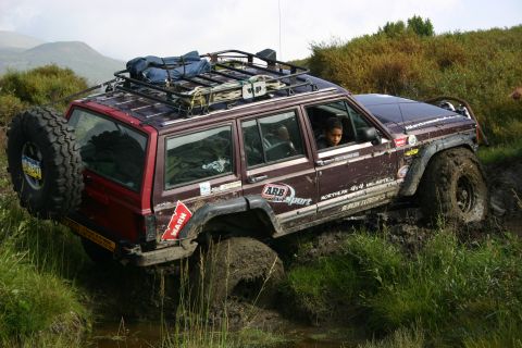 2003 Jeep TJ - A maroon Jeep Cherokee XJ navigates a muddy off-road course, equipped with a roof rack and spare tire.