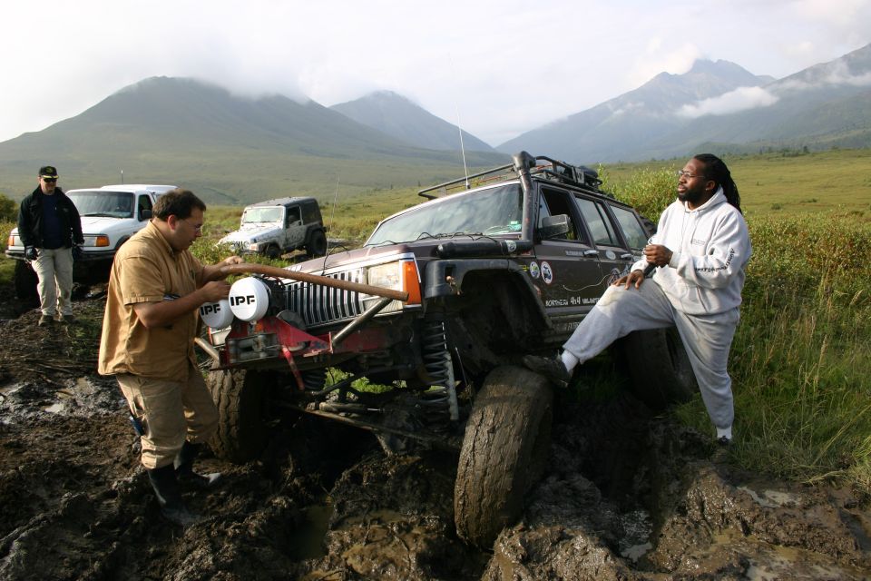 Muddy Jeep Cherokee XJ stuck in a muddy trail with two people nearby; another Jeep and a white SUV are visible in the background