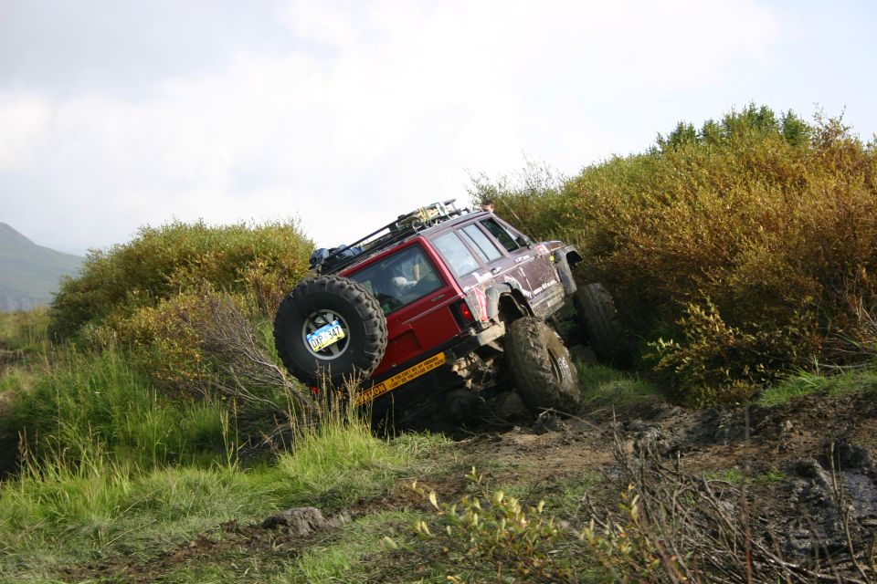 Red Jeep Cherokee XJ with a spare tire on the back, tilted on a muddy hill with green bushes