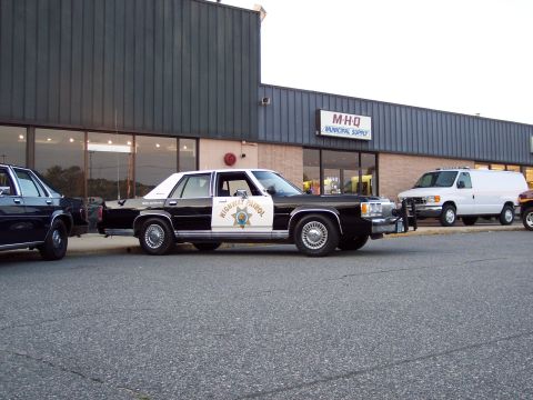 2001 Ford Crown Victoria - A black and white highway patrol car is parked next to a building with a white van behind it.