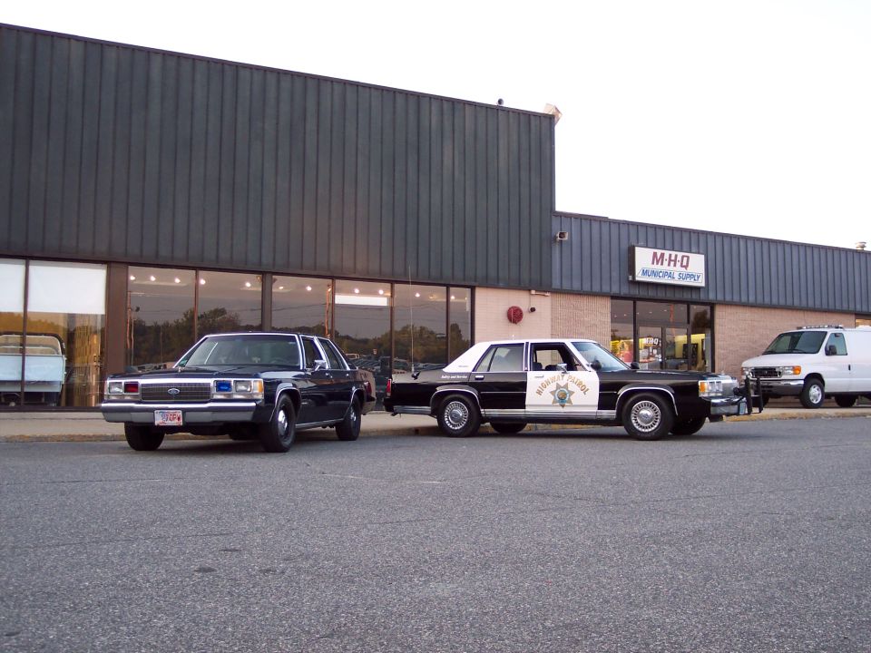 Two sedans parked in front of a building; one is a police car, the other is dark colored. A white van is parked to the right