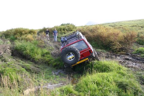 2003 Jeep TJ - A red Jeep Cherokee XJ navigates a steep, muddy off-road trail with two onlookers observing its progress.