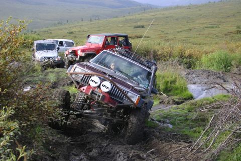 2003 Jeep TJ - Multiple off-road vehicles traverse a challenging muddy terrain with lush green hills in the background.