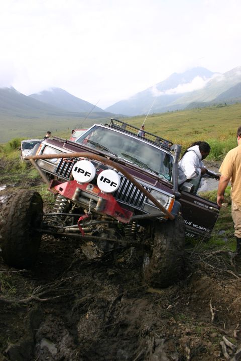 2003 Jeep TJ - A maroon Jeep Cherokee XJ, modified for off-roading, is stuck in thick mud with mountains in the distance.