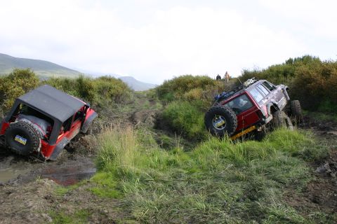 2003 Jeep TJ - Two off-road vehicles struggle through a muddy track on a hillside, with two people visible in the distance.