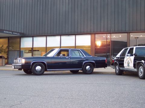 2001 Ford Crown Victoria - A dark blue 2001 Ford Crown Victoria sedan is parked next to a police vehicle outside a building with "MUNICIPAL VEHICLES" signage.