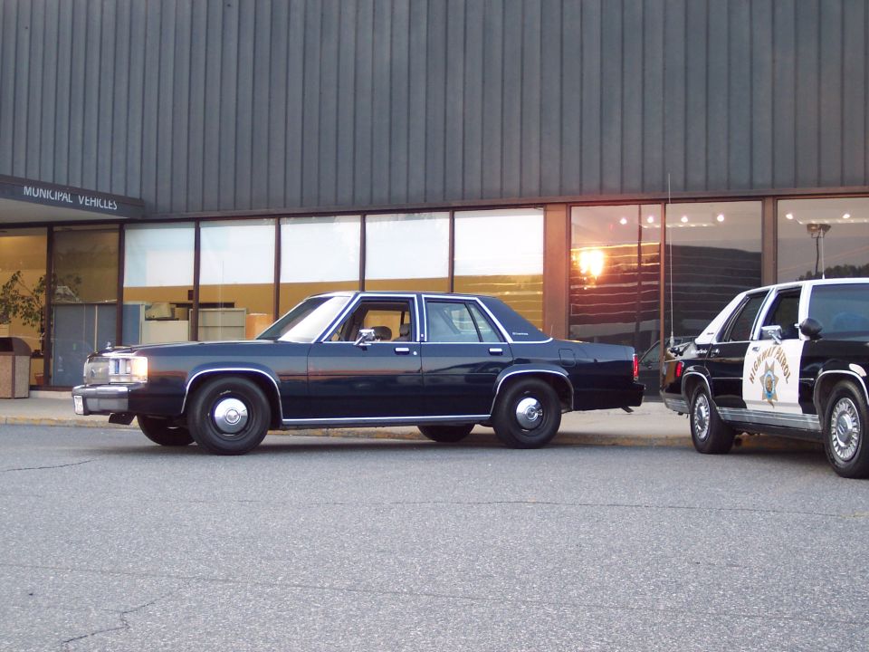 Dark blue 2001 Ford Crown Victoria sedan, side view, parked next to a police car