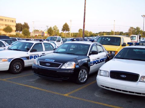 2001 Ford Crown Victoria - Multiple police vehicles are parked in a lot under a bright sky.
