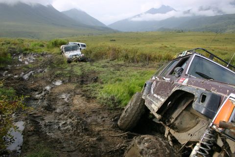 2003 Jeep TJ - Off-road vehicles struggling on a muddy trail with mountainous terrain in the distance.