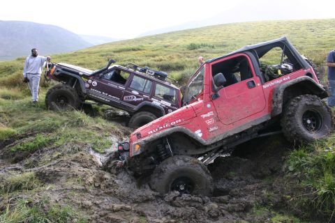 2003 Jeep TJ - Two Jeeps navigate a challenging muddy terrain, one red Wrangler Rubicon is deeply stuck.