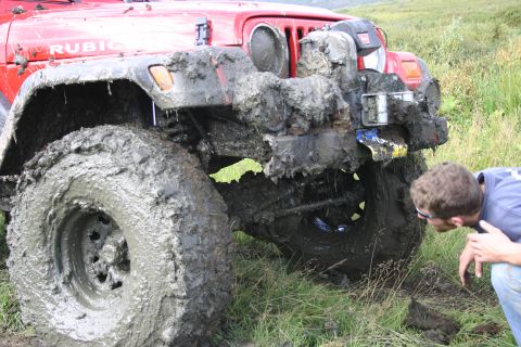 2003 Jeep TJ - A mud-covered red Jeep Wrangler is shown from the front three-quarter view with a person examining its wheel.