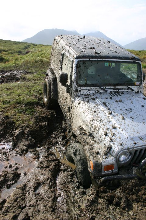 2003 Jeep TJ - A silver Jeep TJ is deeply stuck in thick mud on an off-road trail, with mud splattered across its body and roof.