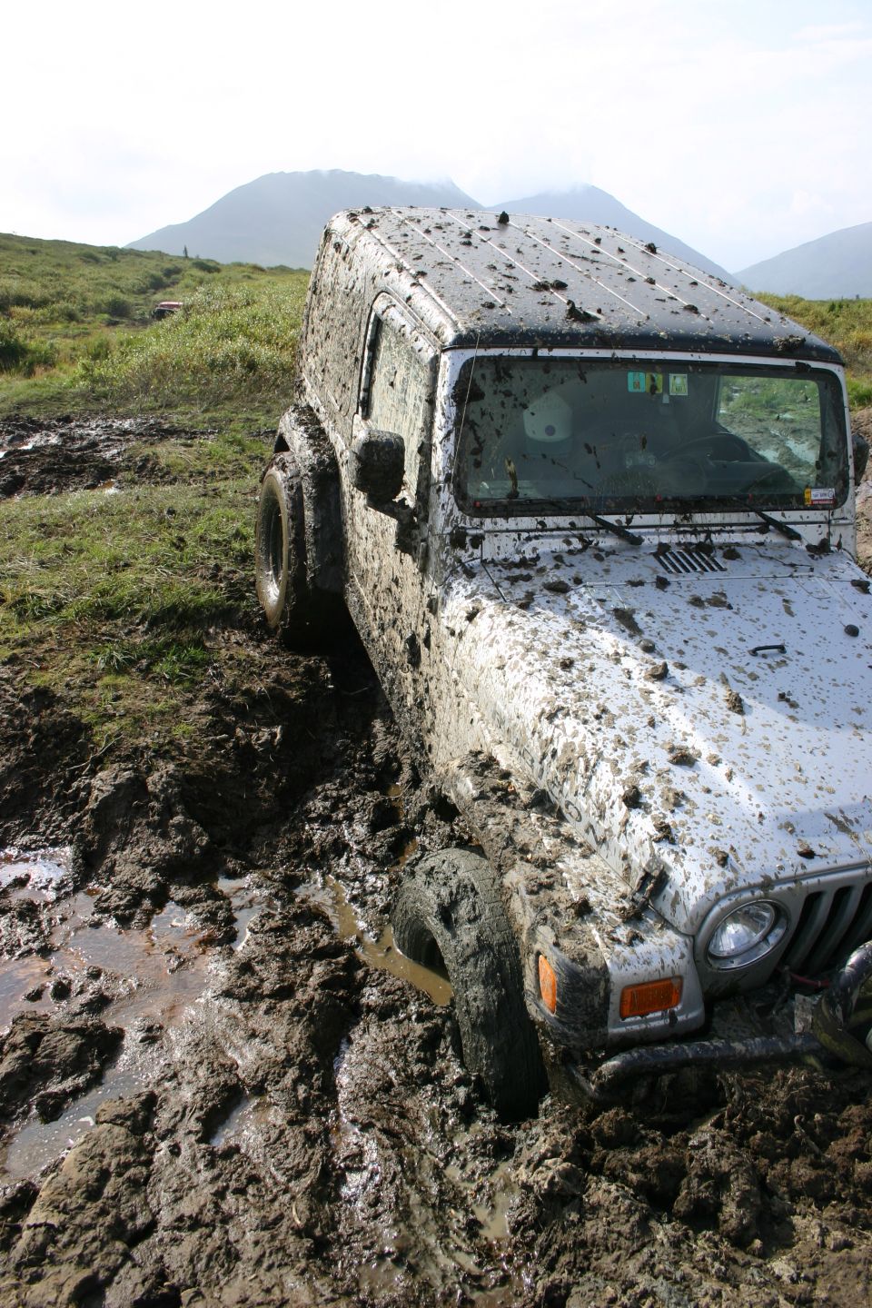 Silver Jeep TJ stuck in mud, front right wheel submerged, covered in mud splatter