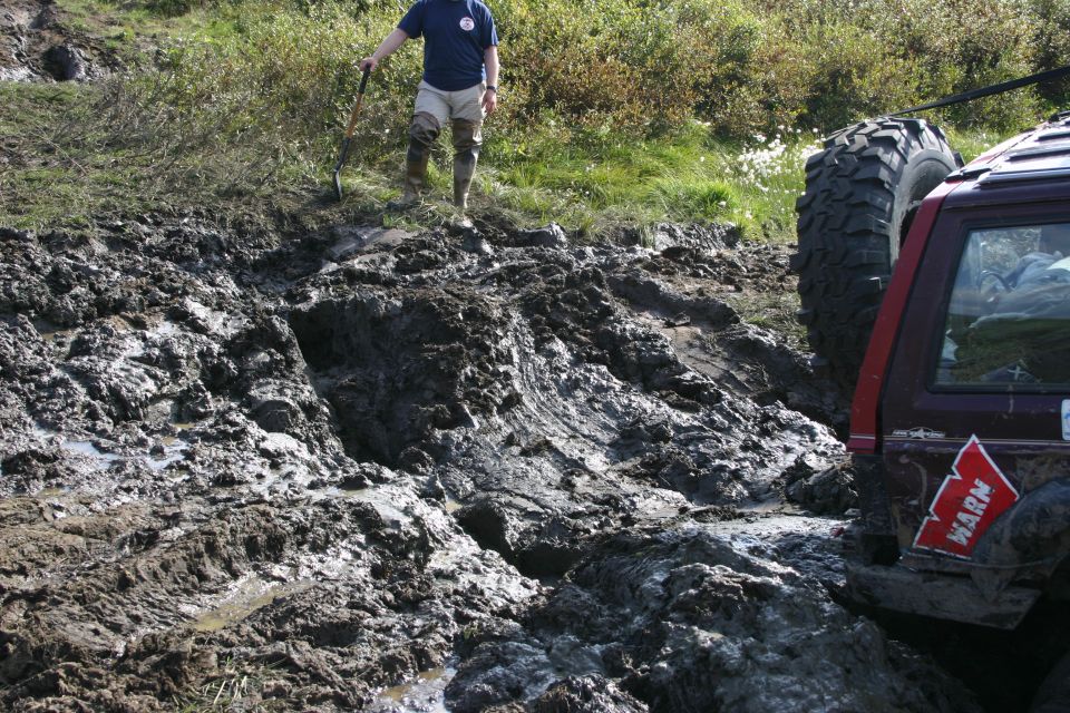 Off-road vehicle with WARN sticker stuck in deep mud, person with shovel standing nearby