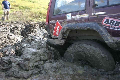 2003 Jeep TJ - A maroon Jeep is deeply mired in thick mud, with one wheel submerged and covered in muck.