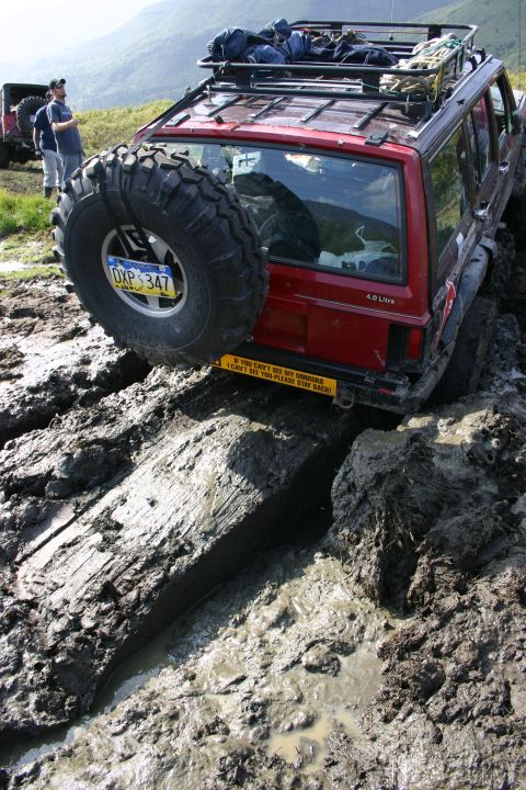 2003 Jeep TJ - A red Jeep Cherokee XJ navigates a muddy trail with a spare tire on the rear and gear on the roof.