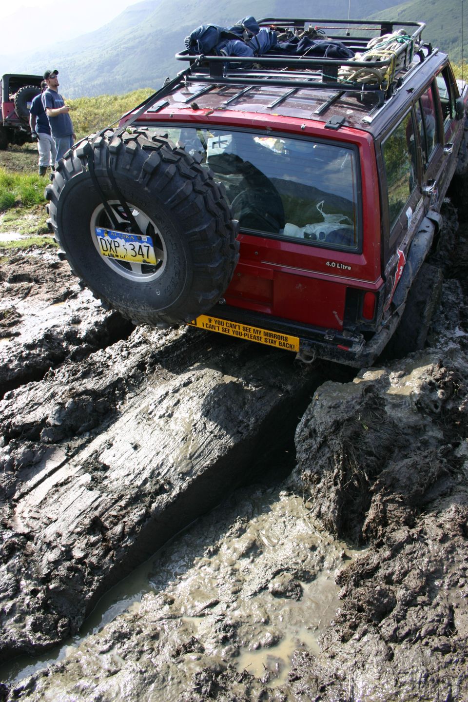 Red Jeep Cherokee XJ stuck in mud, rear view, spare tire mounted on back, roof rack loaded