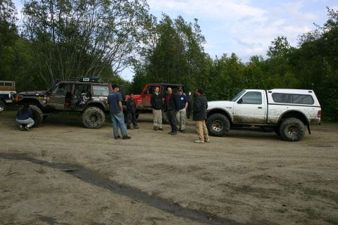 2003 Jeep TJ - Several off-road vehicles and people gather on a dirt lot surrounded by trees.