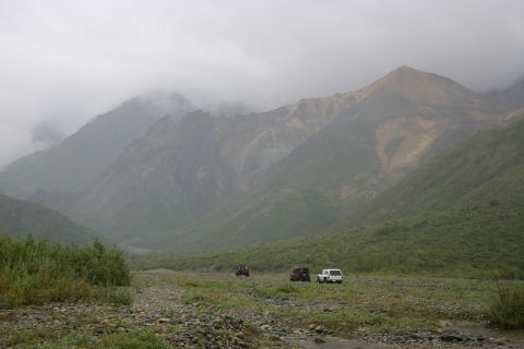 2003 Jeep TJ - Three SUVs traverse a rocky, vegetated valley under a cloudy sky with imposing mountains in the distance.