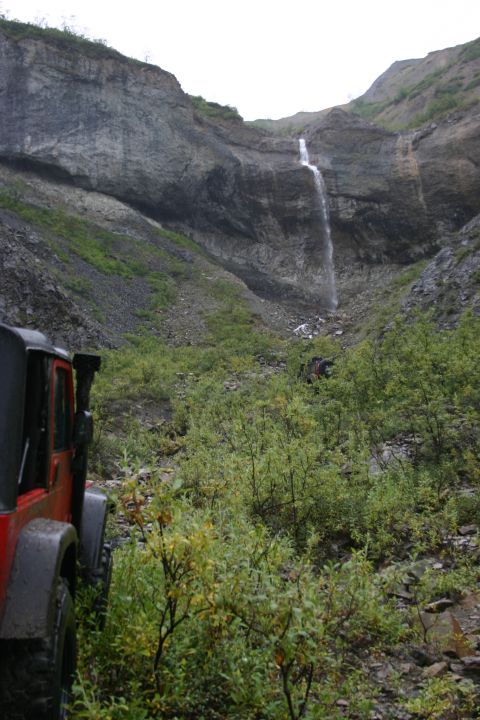 2003 Jeep TJ - A red 2003 Jeep TJ is parked on a , vegetated trail with a tall waterfall cascading down a rocky cliff in the background.