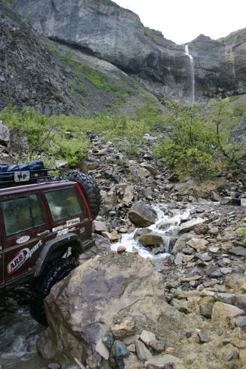 2003 Jeep TJ - A maroon Jeep Cherokee XJ navigates a rocky stream bed with a waterfall cascading down a cliff face in the distance.