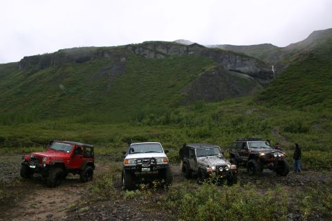 2003 Jeep TJ - Four off-road vehicles are parked on a dirt path in a mountainous, green landscape with a waterfall.