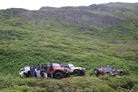 2003 Jeep TJ - A group of off-road vehicles are parked on a vegetated hillside under a rocky cliff face.