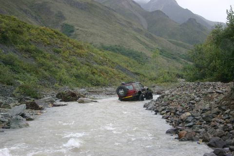 2003 Jeep TJ - A red SUV navigates a rocky riverbed in a mountainous, vegetated area under an overcast sky.