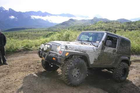 2003 Jeep TJ - A muddy 2003 Jeep Wrangler Rubicon navigates a dirt path, with a scenic mountain range visible behind it.