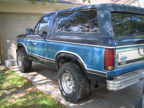 1982 Ford Bronco - A blue 1982 Ford Bronco is parked in front of a garage, showing its rear quarter panel and side profile.