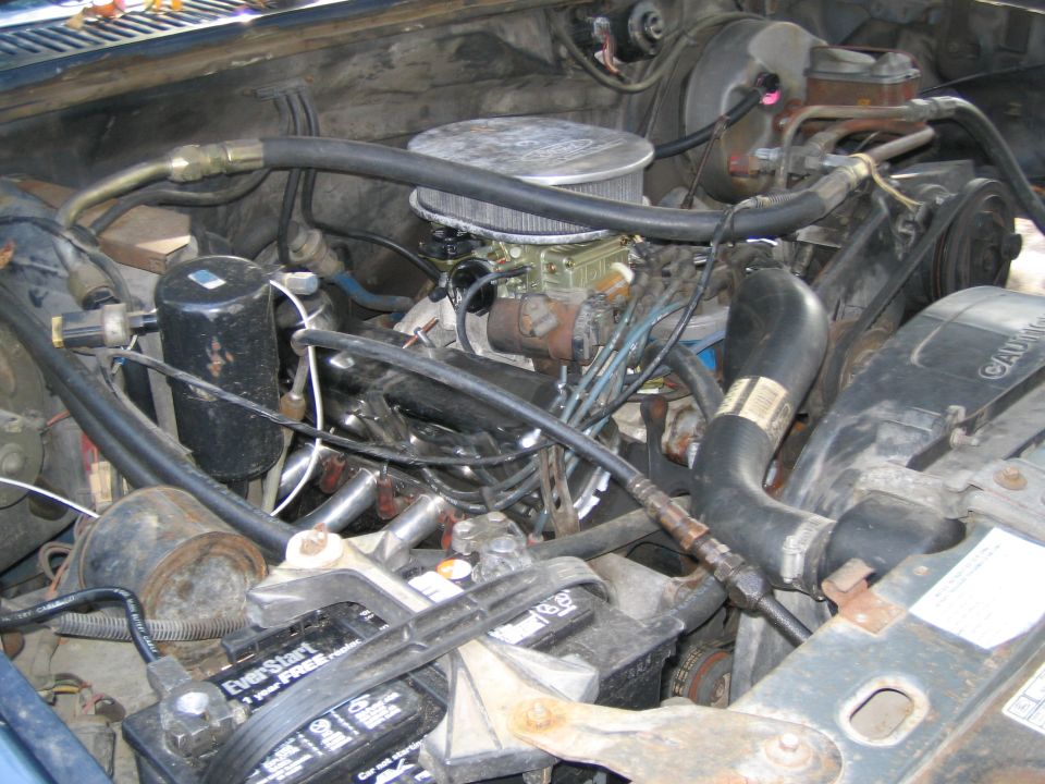 Engine bay with carburetor and battery. Some hoses and wires are visible