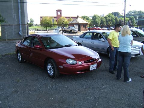 1998 Ford Taurus - A red 1998 Ford Taurus is parked next to other vehicles, with people standing nearby.