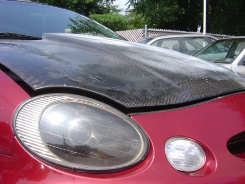 1998 Ford Taurus - A red car with an aftermarket carbon fiber hood and an oval headlight is parked next to another vehicle.